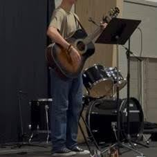 A person stands on stage playing an acoustic guitar and singing into a microphone next to a drum set, with music on a stand, in an indoor performance setting.