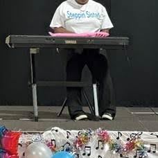 A person wearing a Steppin Sistas t-shirt sits at a Yamaha keyboard on stage, with colorful balloons and musical note decorations in front of the platform.
