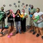 Six adults stand indoors, wearing colorful masks and holding inflatable musical instruments, smiling and posing as if in a band. Vinyl record decorations are on the wall behind them.